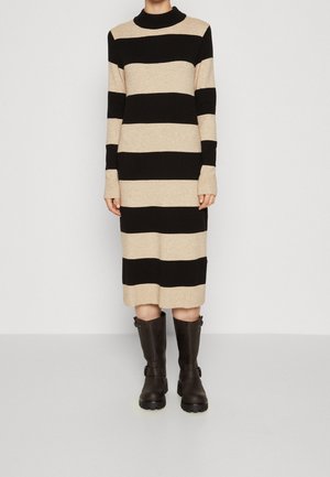 Vrouw draagt een lange, beige en zwarte horizontaal gestreepte trui-jurk met lange mouwen en zwarte halfhoge laarzen met gespen.