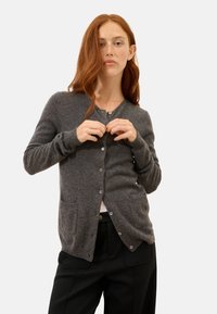 Szary kardigan wykonany z miękkiego materiału, z dekoltem w serek, guziki z przodu oraz dwiema przednimi kieszeniami. Dopasowany krój z długimi rękawami.
