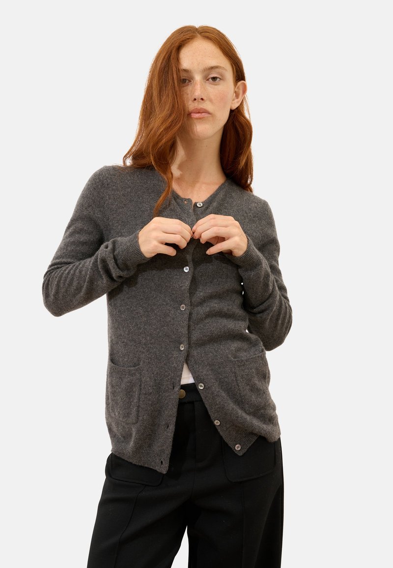 Szary kardigan wykonany z miękkiego materiału, z dekoltem w serek, guziki z przodu oraz dwiema przednimi kieszeniami. Dopasowany krój z długimi rękawami.