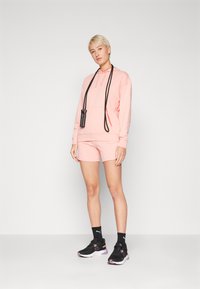 Lys rosa hettegenser med frontlomme og snor, sammen med matchende shorts. Svarte joggesko og et svart hoppetau fullfører antrekket.