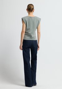 Donna in piedi di spalle, che indossa un top grigio senza maniche con spalline imbottite e jeans a gamba larga blu scuro.