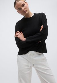 Μαύρο πλεκτό πουλόβερ με ριμπ λαιμόκοψη, άνετη γραμμή και ραμμένους ώμους, συνδυασμένο με λευκό παντελόνι με κορδόνι στη μέση. Απλός και μοντέρνος σχεδιασμός.