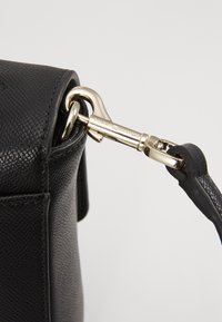 Gros plan sur le coin d'un sac en cuir noir texturé avec un fermoir doré reliant une bandoulière amovible.
