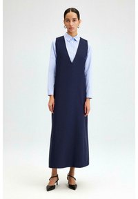 Robe sans manches bleu marine avec un décolleté en V profond, portée par-dessus une chemise à col bleu clair. Design simple, longueur maxi, accompagnée de chaussures à talons noires.
