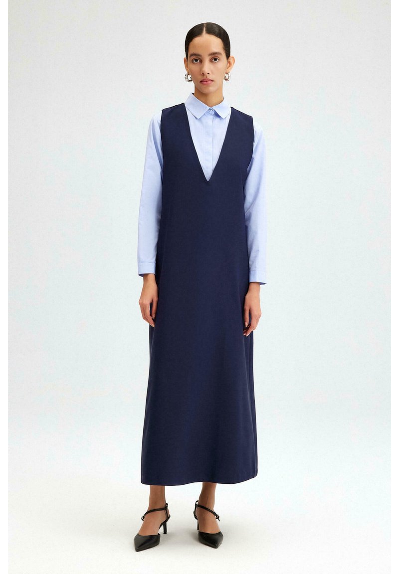 Robe sans manches bleu marine avec un décolleté en V profond, portée par-dessus une chemise à col bleu clair. Design simple, longueur maxi, accompagnée de chaussures à talons noires.