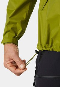 Zielona wodoodporna kurtka z elastycznym pasem, z regulacją za pomocą troczka. Czarne spodnie z boczną kieszenią na zamek i widoczną fakturą materiału.