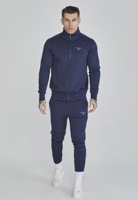Tuta navy con colletto alto e chiusura con zip, realizzata in tessuto testurizzato, design slim-fit e dettagli con logo su giacca e pantaloni.