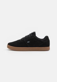 Black suede sneaker with a low-profile design, round toe, black laces, and a rubber gum sole. Side features a subtle logo.