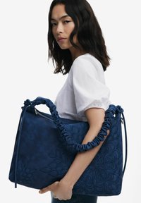 Large blue leather bag featuring a textured floral pattern, gathered handles, and a zip closure, held against a plain background.