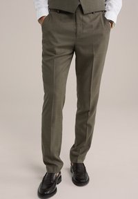 Oliva zöld nadrágok texturált felülettel és sima elülső résszel, oldalsó zsebekkel és szűkített szabással. Fekete cipővel viselve.