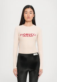 Bodysuit s dugim rukavima u nijansi kože s crvenim logotipom "Fiorucci Cash & Carry"; upareno s crnim sjajnim tajicama, stvarajući elegantnu siluetu.
