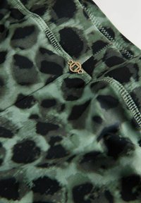 Grønt og sort leopardmønstret stof med glat struktur, prydet med dekorative guldfarvede metaldele og syede sømme. Nærbillede af bærestykkets detalje.