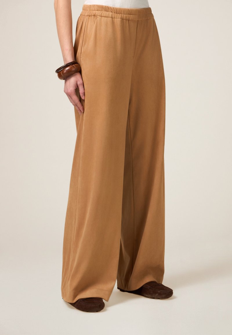 Pantalons larges en tissu couleur brun clair avec une texture douce. Dotés d'une taille élastique et de poches latérales, accompagnés de chaussures brun foncé.