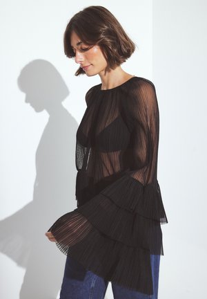 Woman with short brown hair wearing a sheer black layered blouse over a black top and blue jeans, standing against a white wall.