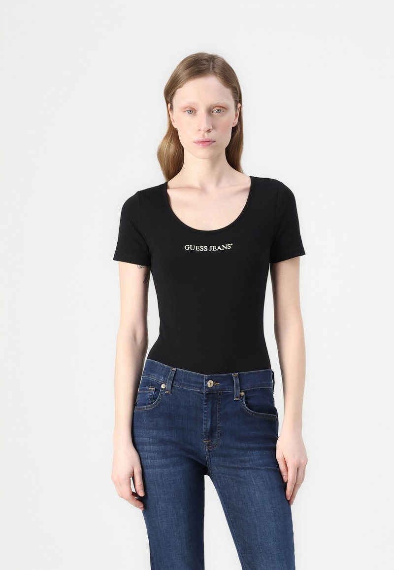 Černý krátký rukáv bodysuit s hladkým výstřihem, zdobený zlatým logem "GUESS JEANS". Kombinace s modrými džíny vytváří kontrast.