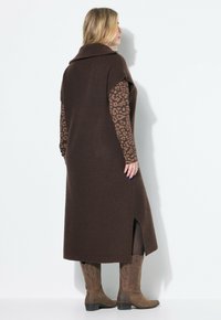 Langer, brauner Strickweste mit breitem Kragen und Seitenschlitzen, kombiniert mit einem braunen, leopardgeprägten Longsleeve; dazu braune Cowboy-Stiefel.