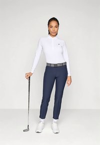 Άσπρο πόλο με μακριά μανίκια και γιακά, σκούρο μπλε παντελόνι στενής γραμμής, μαύρη ζώνη και λευκά αθλητικά παπούτσια. Κλαμπ γκολφ κρατημένο στο αριστερό χέρι.