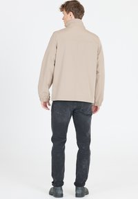 Beige lättviktsjacka med hög krage, elastiska manschetter och avslappnad passform, tillsammans med mörkgrå jeans och svarta ankelkängor.