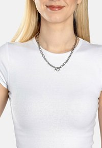 Sølv kædedyse med store led, og en rund lås, båret over en tætsiddende hvid t-shirt, der fremhæver den metalliske tekstur.