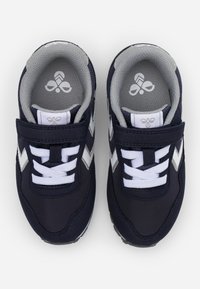 Navy blue sneakers with a smooth fabric upper, white laces, gray interior, and silver accents featuring a chevron design. Velcro strap included.