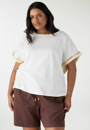 Donna che indossa una maglietta bianca larga con maniche frangiate e pantaloncini marroni con coulisse, su sfondo semplice.