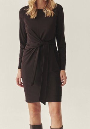 Kobieta ubrana w czarną sukienkę na długi rękaw, długości do kolan, z wiązaniem w talii z przodu oraz czarne kozaki za kolano na jednolitym tle.