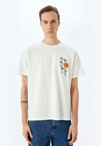 Camiseta blanca de algodón con mangas cortas, con bordado floral en tonos naranjas y verdes en el lado izquierdo. Diseño casual, corte relajado.