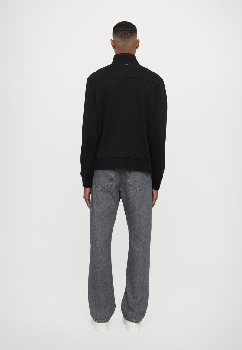 Felpa nera con colletto alto, polsini e orlo a coste. Indossata con jeans grigi larghi e scarpe da ginnastica bianche. Design minimalista.