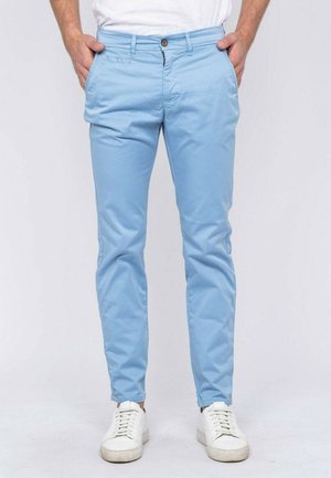 Homme portant un pantalon bleu clair slim et des baskets blanches, les mains dans les poches, debout devant un fond uni.