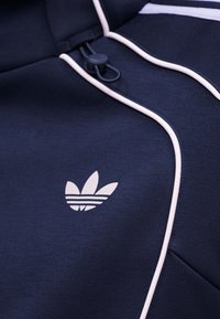 Μπουφάν Adidas σε ναυτικό μπλε με λευκές λεπτομέρειες και κορδόνι στον γιακά, με το λογότυπο Trefoil της Adidas στο στήθος.