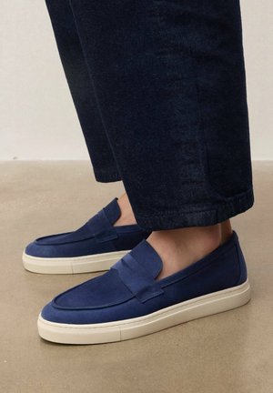 Mocassins bleu marine à enfiler avec semelles en caoutchouc blanches, portés avec un pantalon court bleu foncé sur un sol lisse.