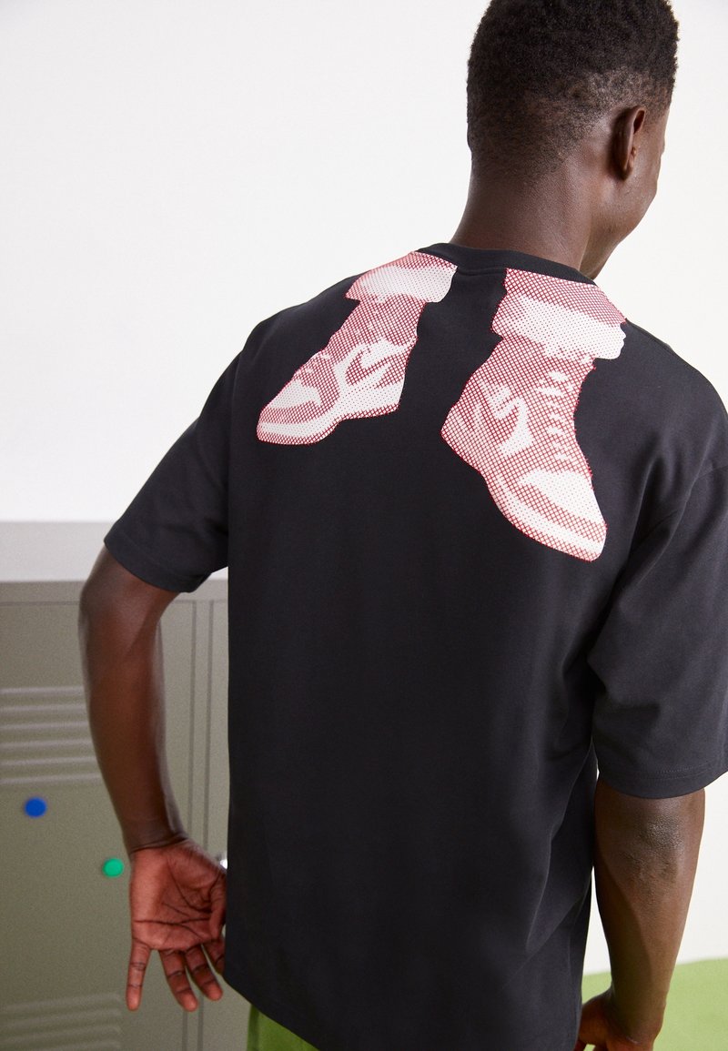 T-shirt noir avec un grand motif de baskets rouges et blanches au dos. Manches courtes, coupe ample et tissu en coton doux.