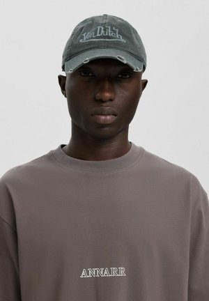 Mladý muž nosící šedou opotřebovanou čepici Von Dutch a taupe mikinu s nápisem "ANNARR" bílými písmeny na hrudi.
