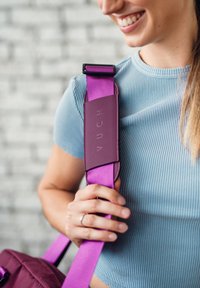 Lila Trage des Bags mit einem strukturierten, gepolsterten lilafarbenen Abschnitt, der mit "VUCH" beschriftet ist. Eine Hand greift die Trage vor einem hellen Hintergrund.