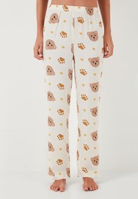 Pantalone za spavanje izrađeni od lagane tkanine, sa ponavljajućim uzorkom lica medvjedice i otisaka šapa u svijetlosmeđoj i žutoj boji na bijeloj pozadini.