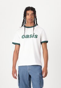 White cotton t-shirt with green ribbed collar and sleeves, featuring bold green "oasis" text on the front, worn with blue cargo shorts.