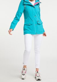 Kvinna som bär en turkos huvtröja, vita uppknytningssmala jeans och vita sneakers med rosa och svarta detaljer, stående på vit bakgrund.