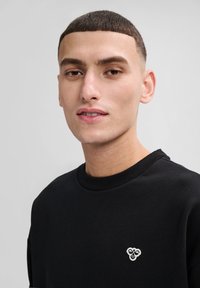 Svart sweatshirt med rund hals, tillverkad av mjukt tyg. Har ett litet vitt emblem på vänster bröst. Slät textur, avslappnad design.