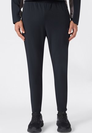Hombre con pantalones casuales negros ajustados y zapatillas negras, de pie con las manos relajadas a los lados, mostrado de la cintura a los pies.