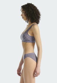 Žena s kudrnatými vlasy v odpovídajícím fialovém vzorovaném podprsenkovém setu, stojící v profilu na jednofonním světlém pozadí.