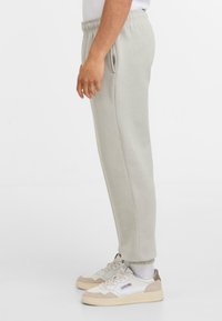 Zijaanzicht van een persoon in lichtgrijze sweatpants met elastische enkelboorden en witte sneakers met beige accenten en logo details.