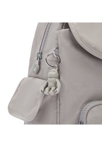 Šedý látkový batoh s hladkým povrchem, který má boční kapsu a zip. Připojen je malý šedý figurkový klíčenka.