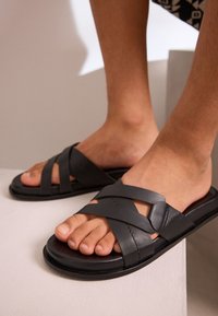 Schwarze Lederschlappen mit einem geflochtenen Riemen-Design und einer flachen Sohle. Die Oberfläche ist glatt mit wenigen Hardware-Details.