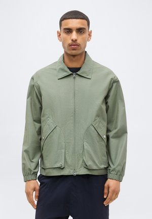 Jeune homme portant une veste zippée vert clair avec de grandes poches devant, debout devant un fond clair uni.