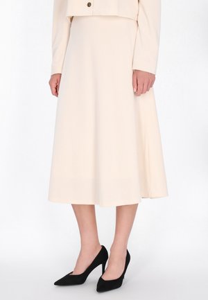 Modelo con una falda midi de color crema y una chaqueta a juego de manga larga, además de zapatos negros de tacón con puntera puntiaguda, sobre un fondo blanco liso.