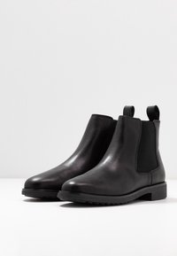 Bottines Chelsea en cuir noir avec panneaux latéraux en élastique, bout rond et semelle en caoutchouc texturé noir. Dispose d'une languette pour un enfilage facile.