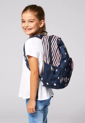 Smiling young girl in white t-shirt and jeans wearing a navy blue backpack with pink stripes and polka dots, facing sideways against a plain background.