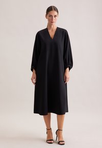 Schwarzes, fließendes Kleid mit V-Ausschnitt und langen, gepufften Ärmeln. Aus glattem Material, knielang mit einem schlichten Design. Kombiniert mit schwarzen Riemchen-Heels.