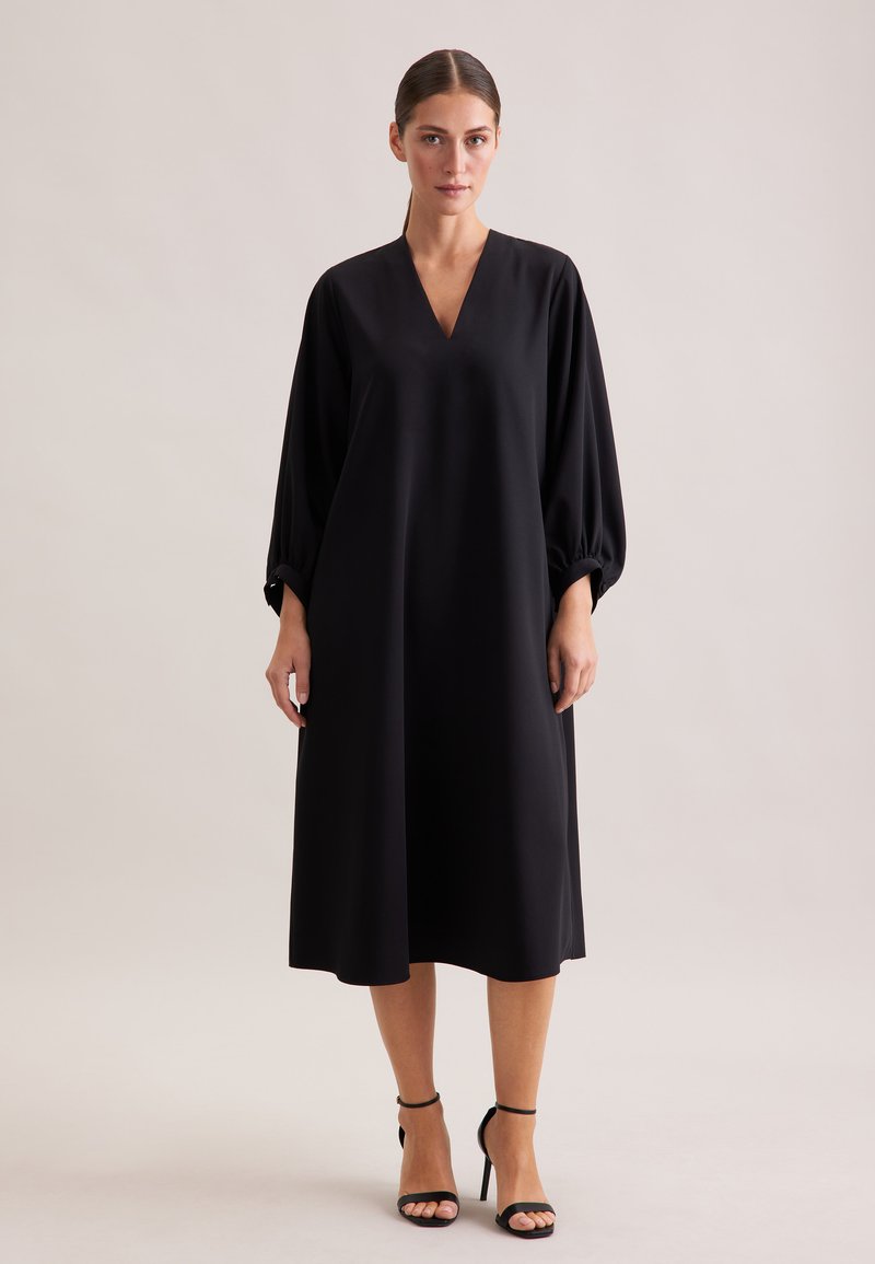 Schwarzes, fließendes Kleid mit V-Ausschnitt und langen, gepufften Ärmeln. Aus glattem Material, knielang mit einem schlichten Design. Kombiniert mit schwarzen Riemchen-Heels.