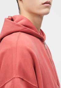 Ροζ φούτερ με ζipper και μεγάλη κουκούλα, με μαλακή υφή και άνετη εφαρμογή. Ορατά ραφές κατά μήκος των ραφών. Χωρίς σχέδια ή διακοσμητικά.